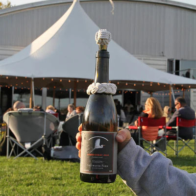 Person holding a wine bottle with a cork in an outdoor setting with tents and people in the background